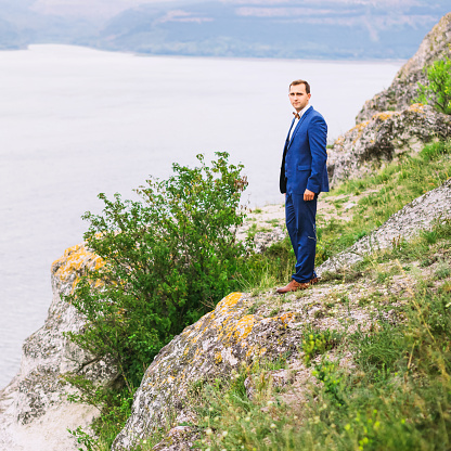 Nice man in blue costume standing at the edge of land with beautiful lake on the background