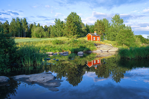 A small red cottage reflected in the river on a beautiful summer evening. An old traditional Finnish sauna cottage.