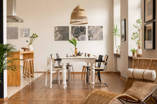 istock Creative composition of dining room interior with mock up poster frame, elegant table, rattan chair, boho lamp, vase with leaves, wooden floor and personal accessories. Home decor. Template. 1645859015