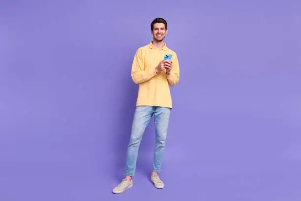 Full length photo of funky cheerful man dressed yellow shirt texting apple samsung iphone device isolated purple color background.