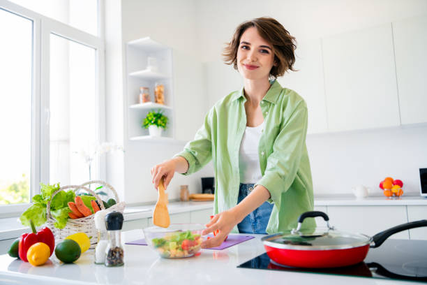 魅力的な女の子の食べ物のブロガーのポートレートは、ヘラミックス野菜サラダボウルフライパン電気ストーブモダンでフラットなインテリアを屋内で保持します - stereotypical housewife women domestic kitchen brown hair ストックフォトと画像