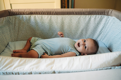 A little baby lying down in his crib next to the parents' bed. He’s smiling.