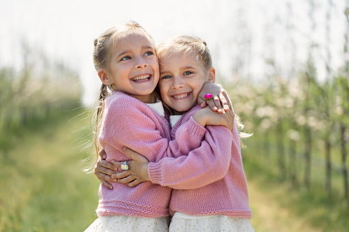 Happy two twin sisters are hugging against the background of a green blossoming apple orchard.