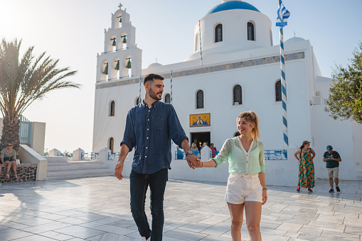 Attractive young travelers holding hands while exploring landmarks of Oia. They are passing by the famous Church of Panagia Platsani. The couple is smiling and looks happy to be spending their vacations traveling around Greece.