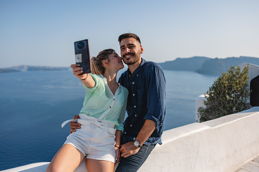 A young tourist couple in Santorini taking a selfie at the viewpoint at the top of a hill. There is a beautiful view of the coast and sea behind the couple. The girlfriend is kissing the cheek of her happy boyfriend. Romantic holiday for couples who love traveling.