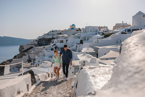 A young adult Caucasian tourist couple happily exploring the beautiful alleys of Oia, Santorini during their summer vacation. The couple is walking uphill.  In the background there is a beautiful view of the iconic white buildings and the sea.