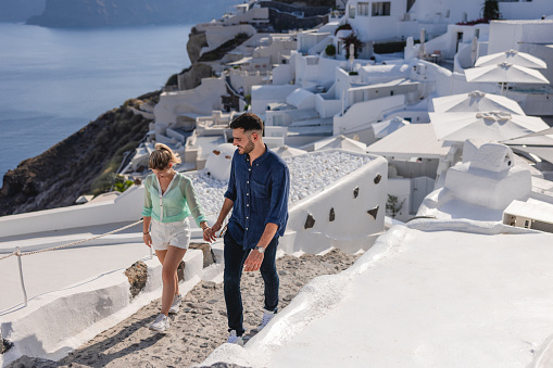 A youthful Caucasian tourist couple walking around the beautiful village of Oia. They are holding hands while walking up some stairs. In The background, there is a beautiful view of the white houses. The couple is having fun travelling and exploring the Greek culture.