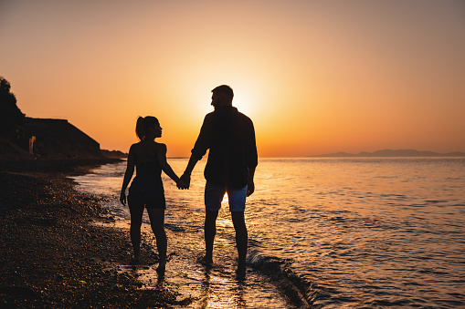 A romantic couple on summer holiday holding hands while walking on the beach in Greece. Only silhouettes are visible. The couple is looking at each other. The sky is golden since the sun is about to set. Romantic beach date in Santorini.