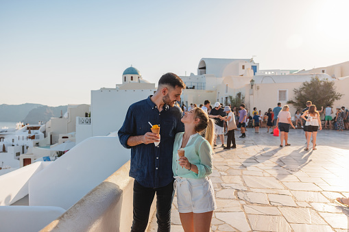 A romantic young adult tourist couple bonding during their vacations in Greece. They are walking around with ice creams in their hands and looking at each other. The sun is setting creating a romantic atmosphere. The couple is dressed nicely and is enjoying their date exploring the village.