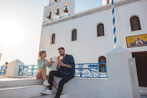 A cheerful tourist couple enjoying their ice cream date by the Church of Panagia Platsani. They are on vacation in Santorini, having a fun time bonding. The couple is sitting by the church facing each other while having a fun conversation. The sun is setting. Romantic summer date in Oia.