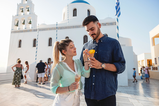 An attractive young adult couple in love sharing an ice cream in Santorini. They are on a date and decided to explore the Island. The couple is standing in front of the Church of Panagia Platsani as the sun is setting. Traveling around Europe and creating memories together as a couple.