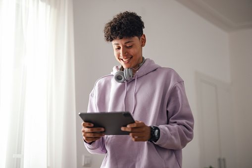 Close up shot of a young smiling boy standing and holding a digital tablet. He is wearing a pink hoodie and  Bluetooth headphones around his neck, smiling while looking down at the screen.