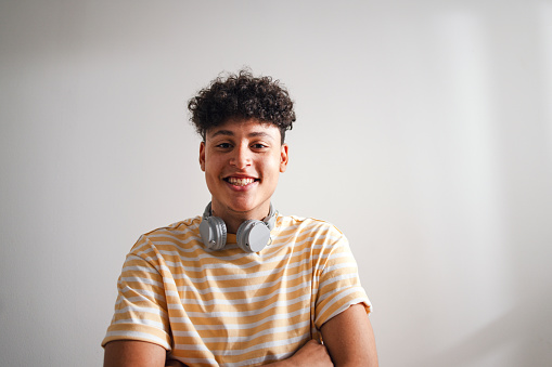 Close up shot of a cute teenage boy in a yellow shirt with Bluetooth headphones around his neck. He is smiling with his arms crossed while looking at the camera.