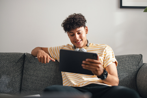 Close up shot of a handsome teenage boy sitting on the sofa at home and using a digital tablet. He is in a yellow t-shirt and smiling while looking down at the screen. There are Bluetooth headphones around his neck.