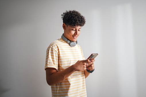 Close up shot of a cute and handsome teenage boy wearing a yellow t-shirt and Bluetooth headphones around his neck. He is standing and using his mobile phone to read or write a text message. He is smiling while looking down at his smart phone.