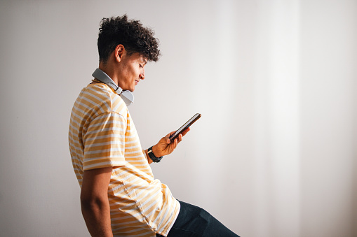 Close up shot of a cute and handsome teenage boy wearing a yellow t-shirt and Bluetooth headphones around his neck. He is sitting and using his mobile phone to read or write a text message. He is smiling while looking down at his smart phone.