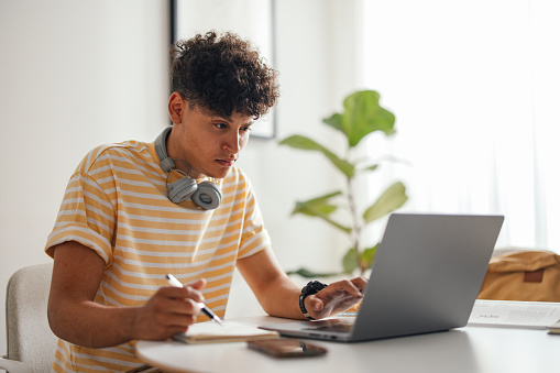 Handsome teenage boy sitting at the table at home doing his homework or studying. He is looking serious and focused while writing notes and using his laptop computer. he is wearing Bluetooth headphones around his neck and a big watch.