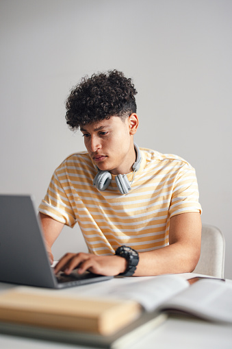 Handsome teenage boy sitting at the table at home doing his homework or studying. He is looking serious and focused while using his laptop computer. he is wearing Bluetooth headphones around his neck and a big watch.