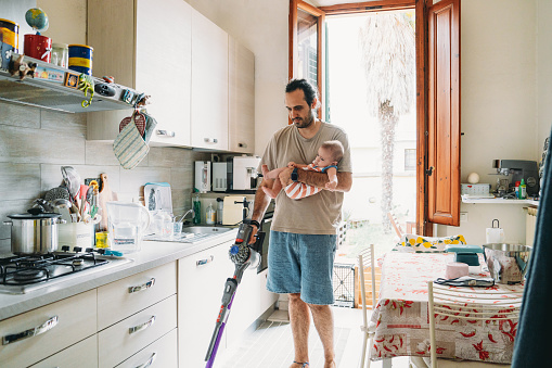 A father is using vacuum while he's holding his son. Modern fatherhood concept.