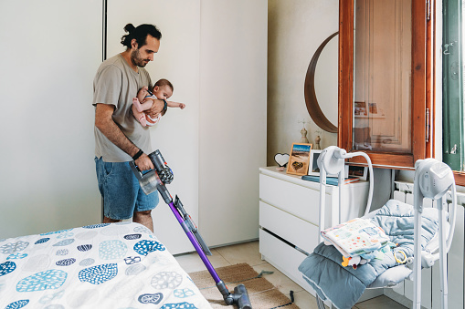 A father is using vacuum while he's holding his son. Modern fatherhood concept.