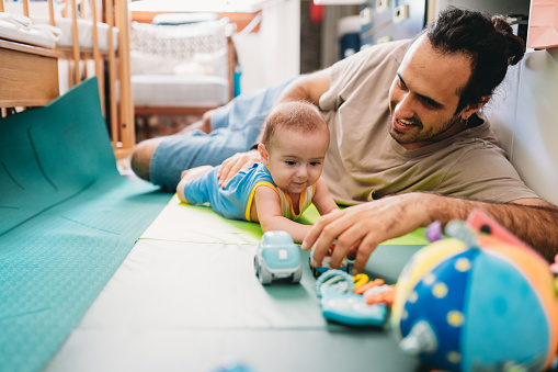 A father is playing with his son at home. They are having fun together. The baby is 5 months old and he's doing tummy time.