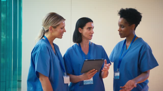 Three Mature Female Doctors Wearing Scrubs With Digital Tablet Discussing Patient Notes In Hospital