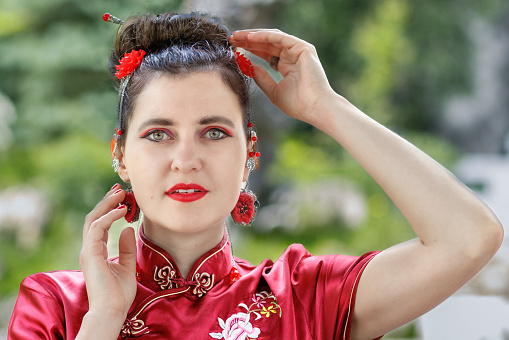 A young Asian girl walks in national clothes with a fan in the park. Chinese woman in a red traditional dress, close-up portrait.