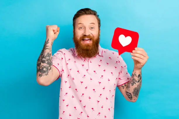 Photo of Portrait of lucky cheerful man with cool tattoo wear flamingo t-shirt hold like icon support you isolated on blue color background