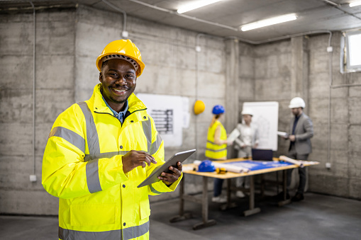 Construction site workman in safety work wear holding tablet computer while his coworkers having a meeting in background.