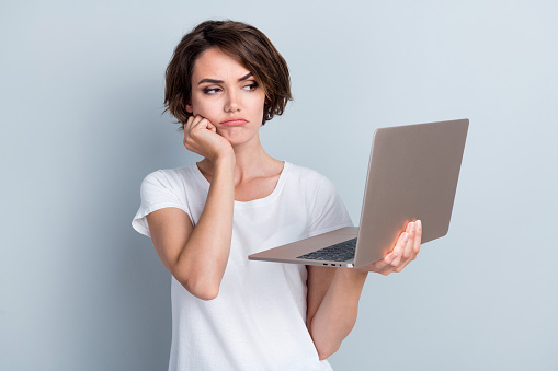 Photo of disappointed girl dressed white t-shirt holding laptop fist on cheekbone watch boring video isolated on gray color background.