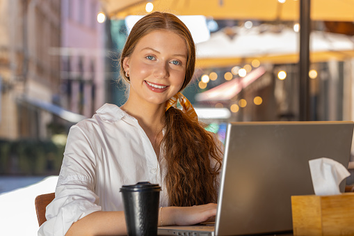 Redhead child girl working study education online distant job with laptop in city sunny street cafe restaurant browsing website chatting outdoors during break. Smiling woman tourist looking at camera