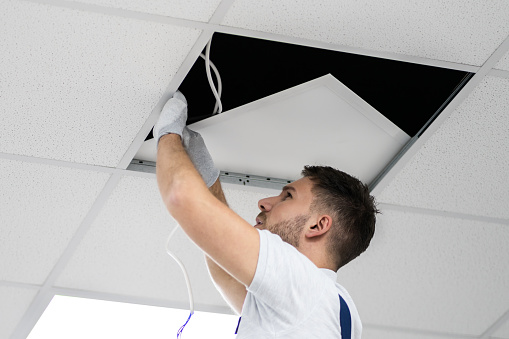 Full Length Portrait Of Electrician On Stepladder Installs Lighting To The Ceiling In Office