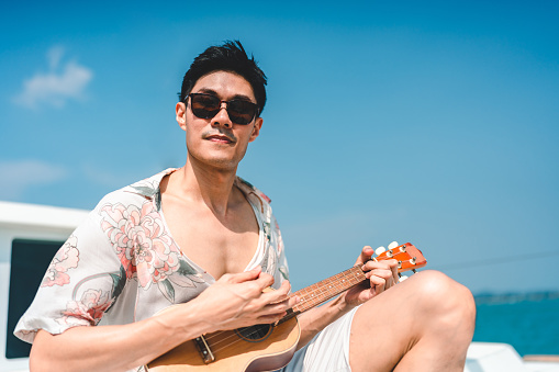 Outdoor travel lifestyle concept, Asian man playing ukulele or guitar music in holiday vacation, Male having fun on sailboat or yacht boat on the sea ocean at sunset, cruise ship at tropical island