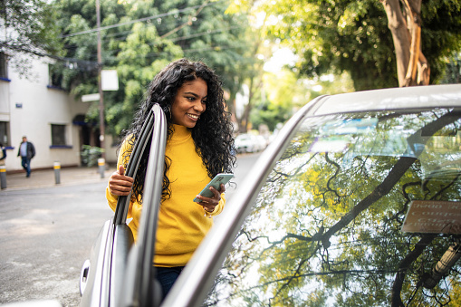 A young woman standing on the street and opens a car door. She uses a mobile phone.
