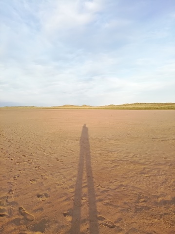 Long shadow of a person on beach at sunset with grassy dunes in background