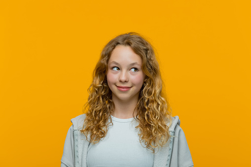 Curly blond hair teenage girl wearing grey hoodie and white top looking at copy space and smiling. Studio shot, yellow background.