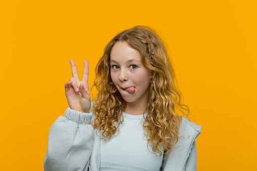 Curly blond hair teenage girl wearing grey hoodie looking at camera, showing peace sign and sticking out tongue. Studio shot, yellow background.