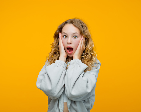 Shocked curly blond hair teenage girl wearing grey hoodie holding face in hands and looking at camera with mouth open. Studio shot, yellow background.