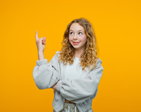 Curly blond hair teenage girl wearing grey hoodie pointing with index finger at copy space, looking up and smiling. Studio shot, yellow background.