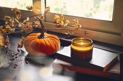 Cozy autumn still life with pumpkin, books and burning candle on the windowsill. Autumn home decor. Cozy fall mood.