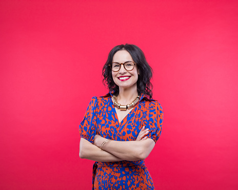 Happy mature woman wearing smart casual dress standing with arms crossed and smiling at camera. Studio shot, red background.