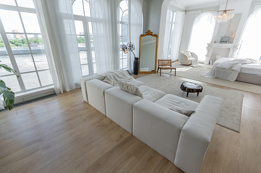 cozy bright apartment with huge panoramic windows with sunlight. bedroom in white and beige tones with fireplace and soft armchair. stylish living room with log coffee table and large white sofa