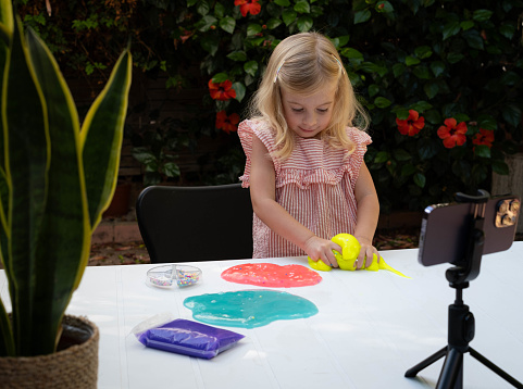 Cute blonde caucasian girl with curly hair playing with slime and watching video lesson on smart phone. Online instruction of preparing concept. Kid sensory development, creative hobby, game.