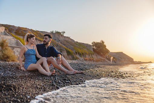 A young adult Caucasian couple happy to be spending their holidays together at such a romantic destination as Santorini. They are sitting on a beautiful beach, talking and enjoying their time together. The sun is setting and creating a nice and romantic atmosphere. Romantic summer date ideas for creating new memories.