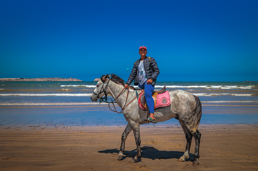 Essaouira, Morocco - August 3, 2023: A boy on horseback looking for tourists on the beautiful beach overlooking the atlantic ocean.