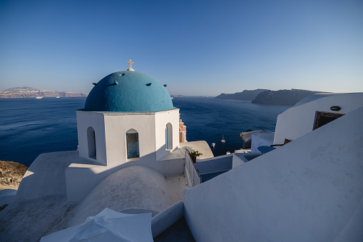 A beautiful view of the iconic blue and white houses and the sea in Santorini. The sun is partially illuminating the village. The sea is calm and blue. Beautiful and luxurious travel destinations.