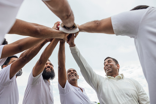 Directly below view of male friends and family members ready to play cricket together. They are wearing white standing with their hands together in a huddle on a sunny day in Northumberland.