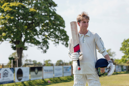 Young male cricket player walking onto the pitch holding a cricket bat and helmet ready to play for his team. It's a sunny day in Northumberland.