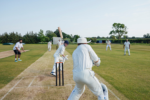 Male batsman hitting ball on cricket pitch while wicketkeeper standing behind stumps.