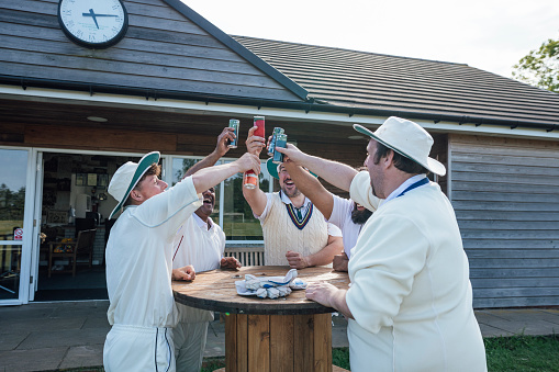 Male cricket team members celebrating together after playing cricket. They are wearing white standing with their hands together, toasting with their soft drinks around a table on a sunny day in Northumberland.